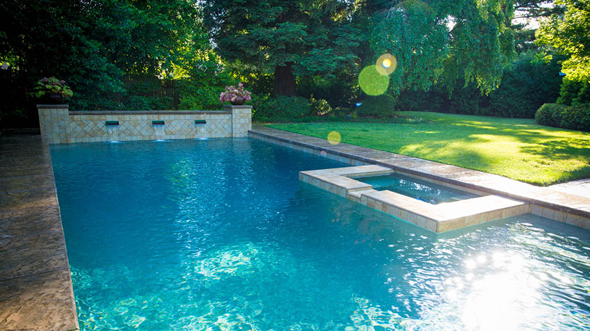 Rectangular outdoor swimming pool with clear water, built-in water features, and surrounding green lawn and trees on a sunny day.