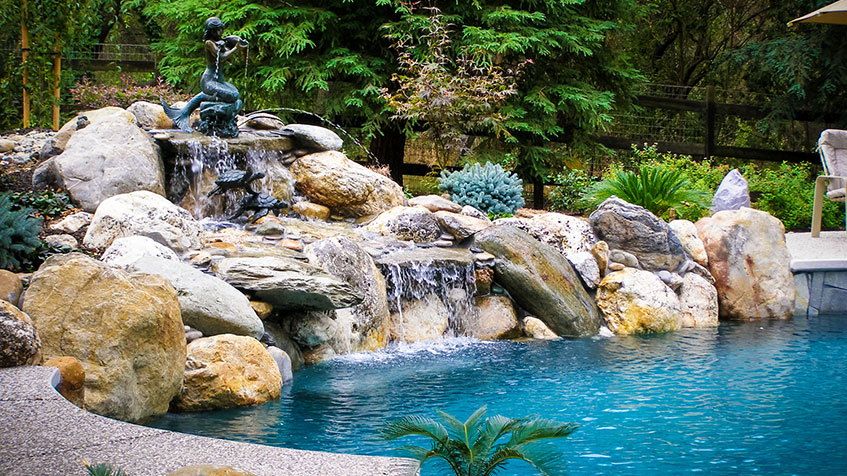 A natural-style pool with rock waterfalls and a bronze mermaid statue surrounded by lush greenery.