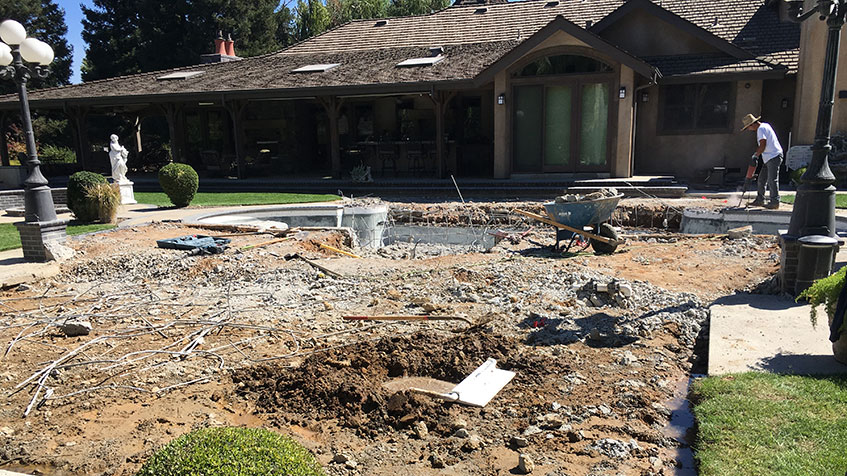 Construction site in front of single-story house showing excavated courtyard area with exposed dirt, broken concrete, construction debris, and rebar. Worker visible in background near house entrance, white statue and landscaping visible on intact lawn areas.