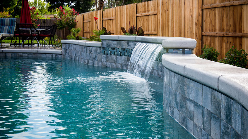 A backyard pool features a smooth stone wall with a narrow waterfall and patio seating nearby.