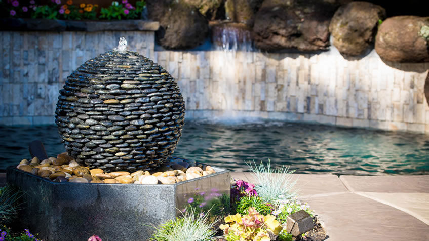 A round stone fountain made of stacked pebbles sits by a pool with flowers at its base.