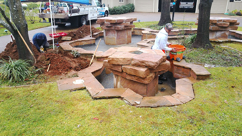 Workers install a stone water feature in a grassy yard with construction tools and equipment nearby.
