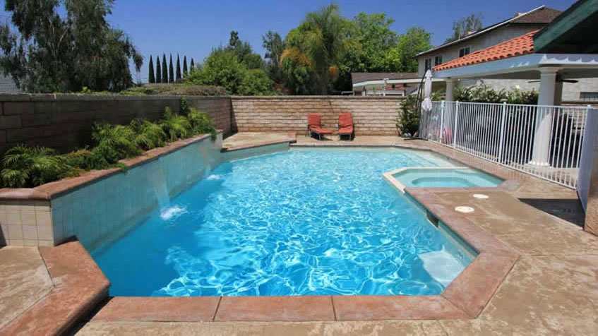 A backyard pool with a built-in spa and small waterfalls flows beneath a sunny blue sky.