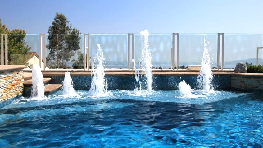 Jets of water shoot up from a modern pool with glass fencing and a view of distant hills.