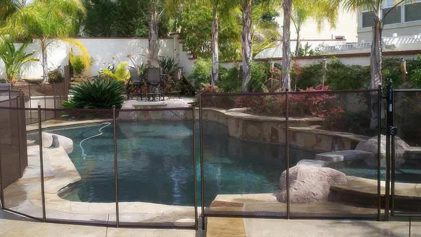 A fenced backyard pool is surrounded by palm trees, stone decking, and tropical landscaping.