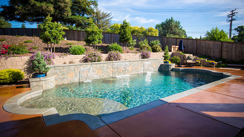 A finished backyard pool with clear blue water, waterfalls, and surrounding landscaping.