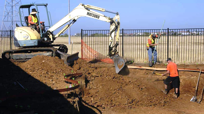 Construction workers dig a pool area using a small excavator and hand tools.