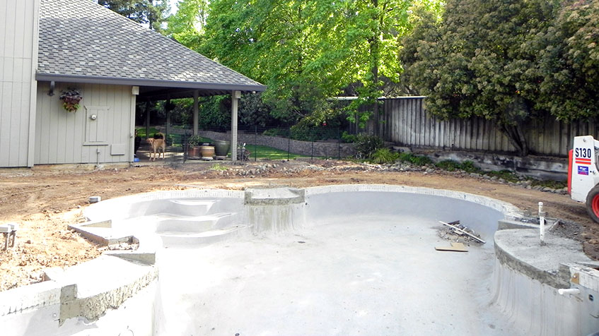 A shaped concrete pool shell sits unfinished beside a house in a dirt yard.