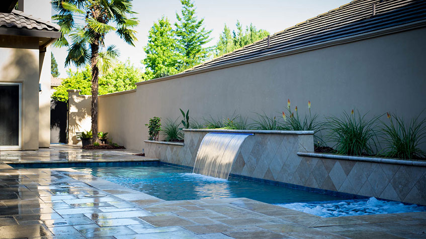 A narrow backyard pool with a tiled waterfall feature and tropical landscaping along a privacy wall.