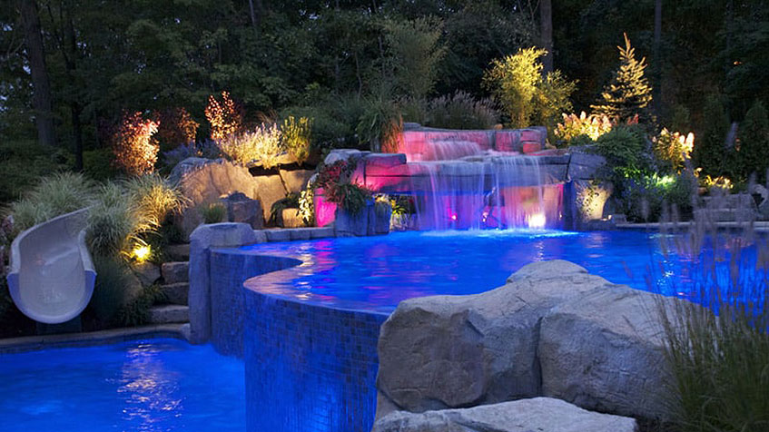 A glowing infinity-edge pool with a lit-up rock waterfall and lush landscape at night.