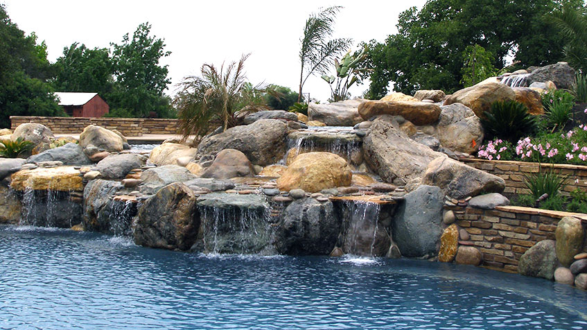 A resort-style pool bordered by a large, tiered rock waterfall with tropical plants.
