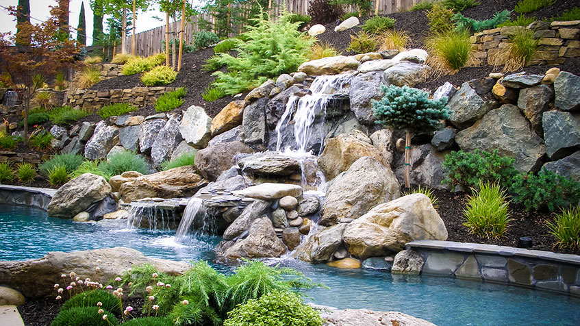 Multi-tiered rock waterfall feature flowing into swimming pool, constructed with large natural boulders and stones, surrounded by extensive landscaping including ornamental grasses, evergreen shrubs, and perennials on terraced hillside with stone retaining walls.