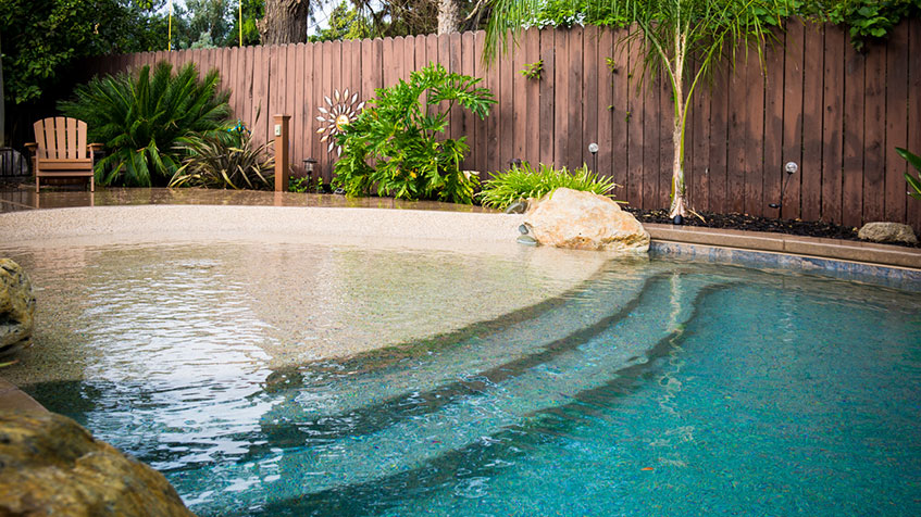 A shallow entry pool with a beach-style slope and tropical plants beside a wooden fence.