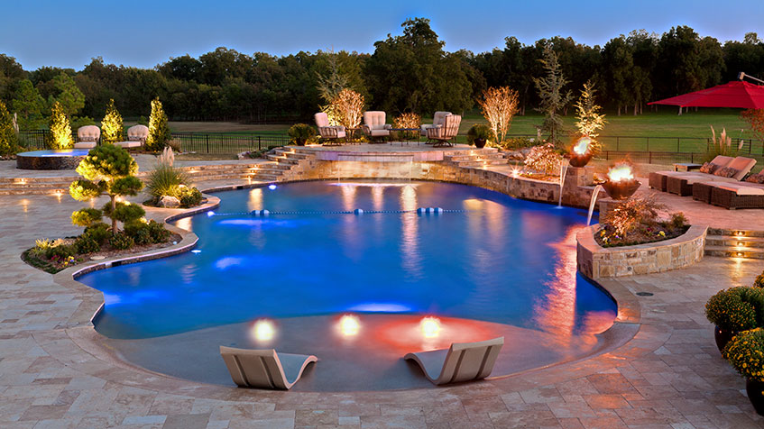 A large luxury pool glows with underwater lights and fire features, surrounded by trees and lounge chairs.