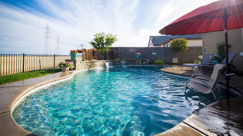 A sunny backyard pool with curved edges, a red umbrella, and small waterfalls near a wooden fence.