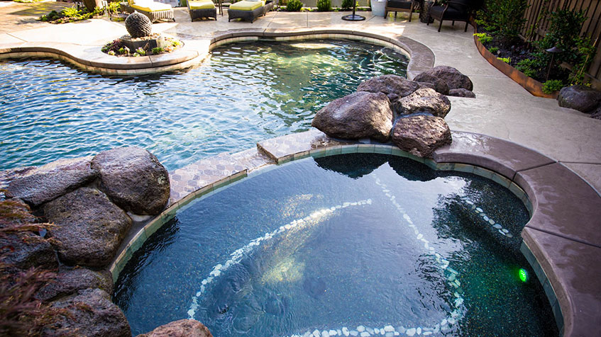 A multi-level backyard pool with a round spa in the foreground and smooth stone accents.
