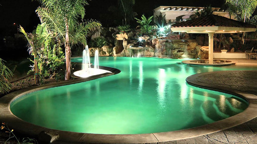 A curvy backyard pool at night illuminated with green lighting and surrounded by palm trees.