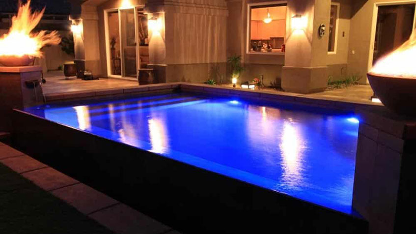 A glowing blue rectangular pool at night with two fire bowls and patio lighting.