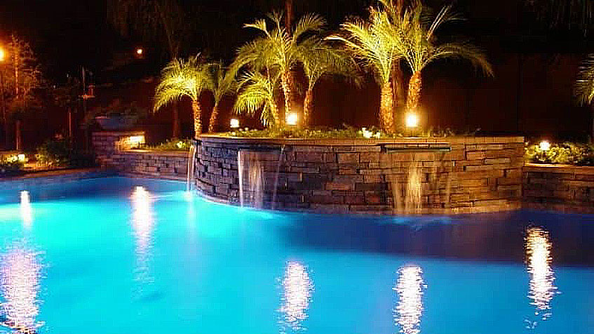 A night-lit pool with stone walls, cascading waterfalls, and palm trees reflecting on the water.