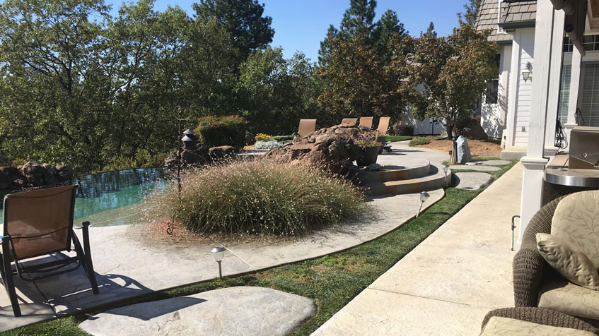 A hillside infinity-edge pool beside a house is framed by patio furniture and tall grasses.