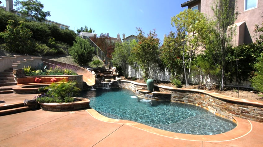 A curvy hillside pool with a built-in fountain and waterslide is surrounded by terraced landscaping.