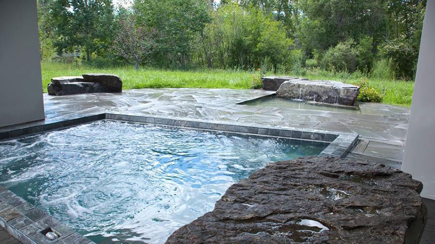 A square spa with dark stone tiles sits on a flagstone patio facing a natural grassy landscape.