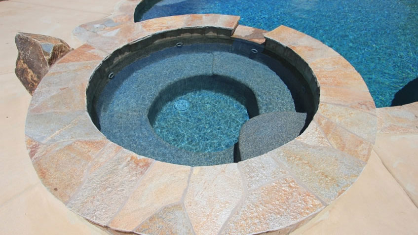A circular stone spa with steps is built into a pool deck next to clear blue water.
