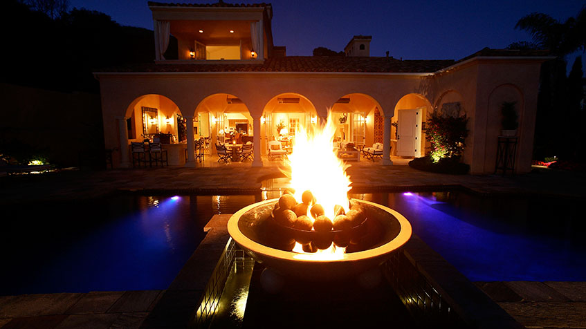 A luxurious night-lit backyard with a glowing fire bowl centered over a narrow pool.