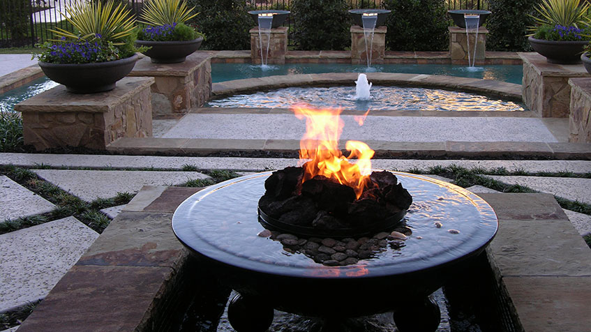 A modern fire bowl sits in front of a round spa with fountains and symmetrical landscaping.