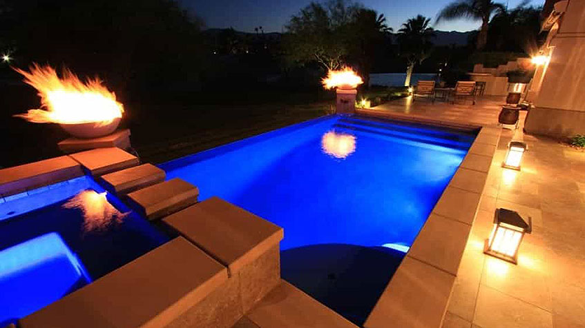Nighttime view of illuminated rectangular swimming pool and spa with bright blue underwater lighting, concrete stepping stones connecting pool to spa, fire bowls with active flames on both sides, decorative pathway lights along pool edge, lounge chairs on patio, palm trees and tropical landscaping in background at dusk.