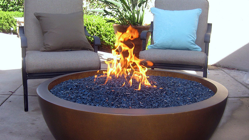 A fire pit with blue fire glass sits between two cushioned chairs on a backyard patio.