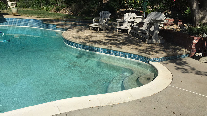 A curved pool with turquoise tile trim and a deck area featuring Adirondack chairs.