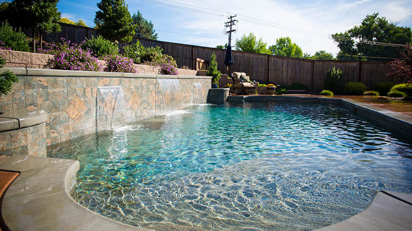 Swimming pool with curved shallow entry area featuring multiple water jets creating fountain effects, stone veneer wall with waterfall spillways, clear blue water, concrete decking, landscaped raised planter with purple flowering plants and shrubs, wooden privacy fence, and utility poles visible in background.