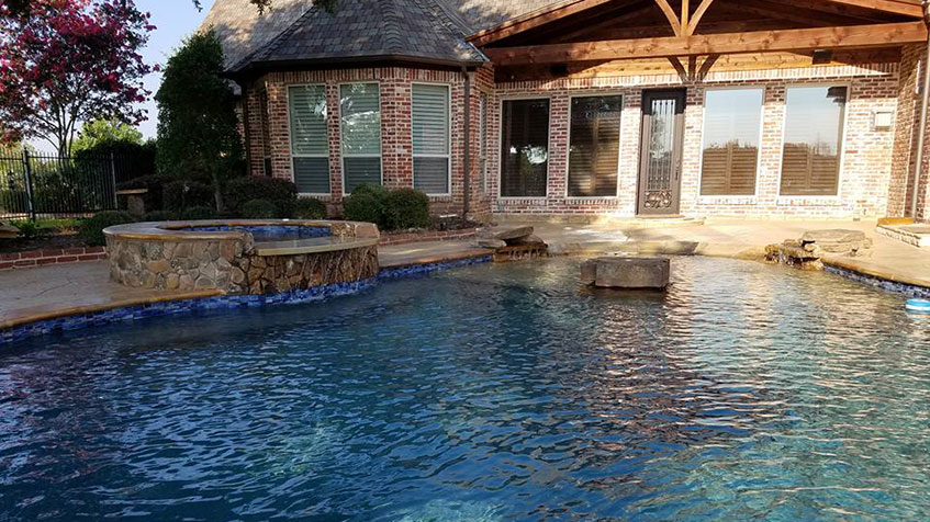 A lagoon-style pool with a raised stone spa sits in front of a brick home with large windows.