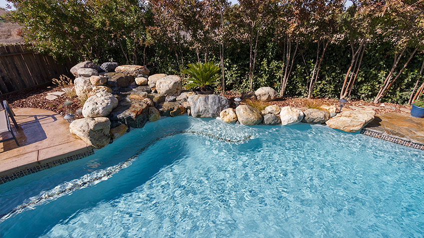 A bright blue backyard pool features a shallow tanning ledge beside a rock waterfall and trees.