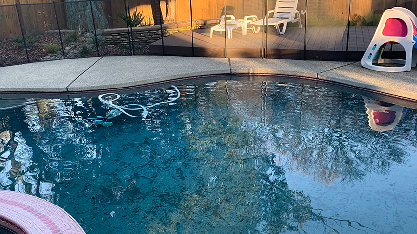 A slightly dirty pool with a cleaning hose and mesh safety fence near a child’s play area.