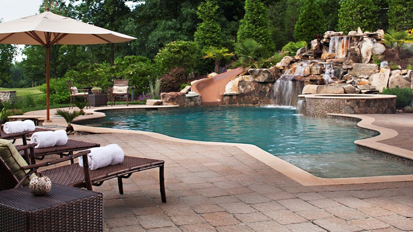 A resort-style pool with a waterfall, lounge chairs, and a curving waterslide surrounded by landscaping.