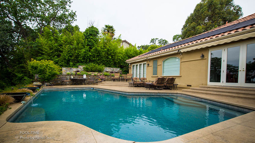 A rectangular backyard pool beside a stucco home with an outdoor dining patio and waterfall