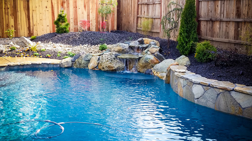 A backyard pool with a small rock waterfall and fresh landscaping along a wooden fence.