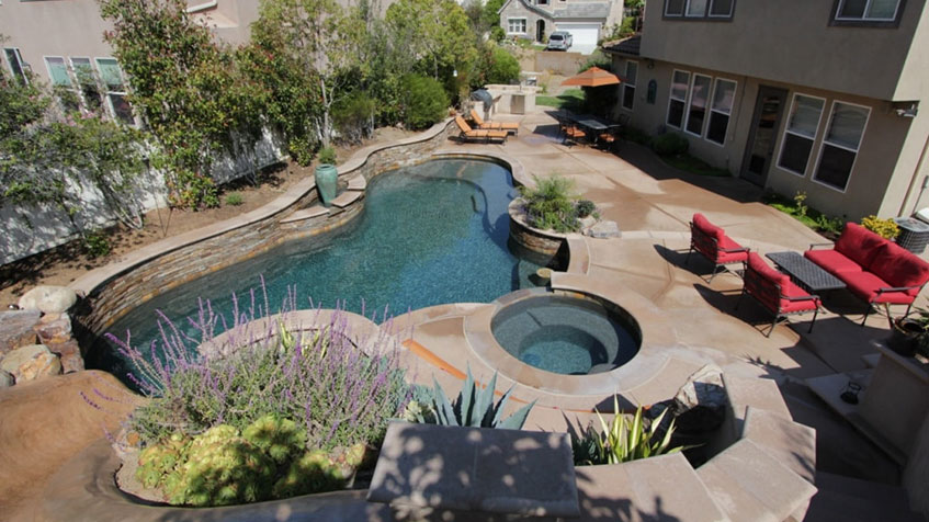 A curvy backyard pool with a spa, stone features, and patio seating wraps around a two-story house.