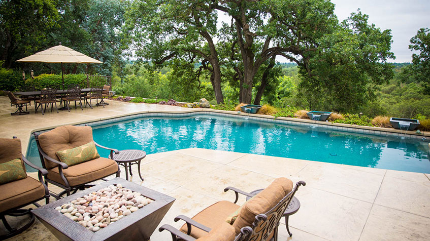 A rectangular pool with a stone patio and lounge seating overlooks a wooded landscape.