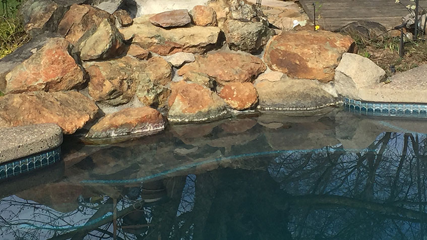 A pool bordered by rough rocks with calm water reflecting the trees above.