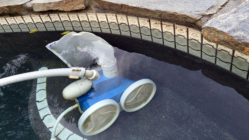 A pool cleaning robot partially submerged in water, resting near a decorative tile border.