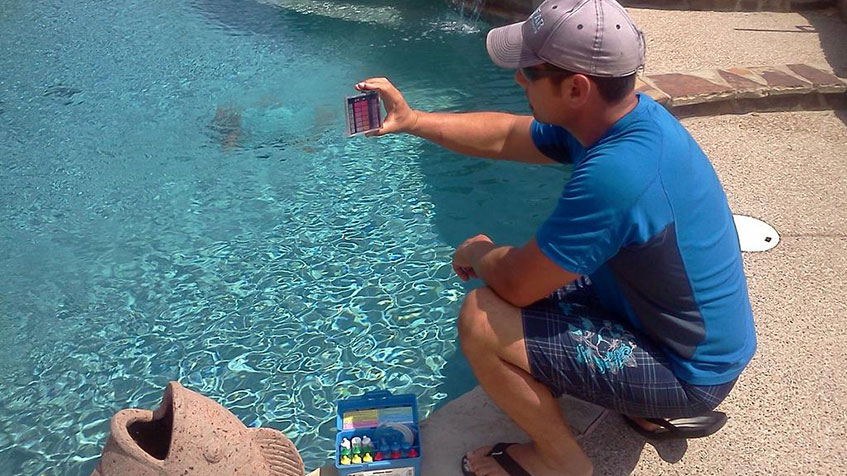 A man kneels at the edge of a pool while testing water quality using a test kit.
