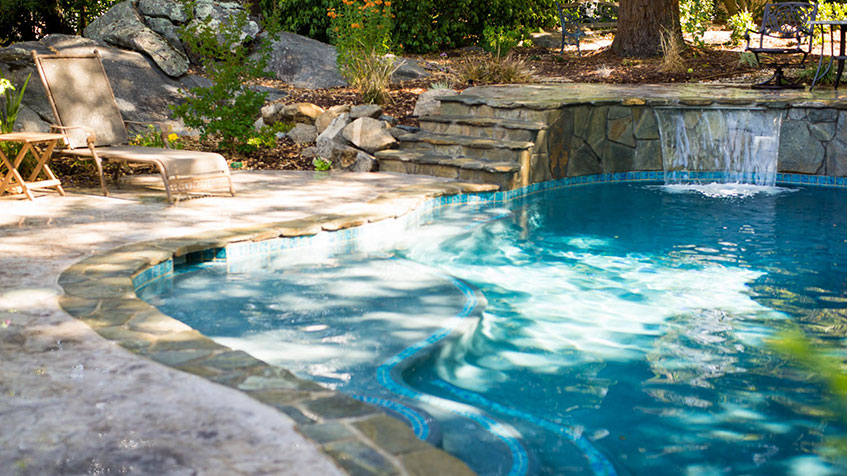 An outdoor swimming pool with blue water featuring a stone waterfall spillway, surrounded by natural landscaping with rocks and plants, and a beige lounge chair on the stone patio deck.