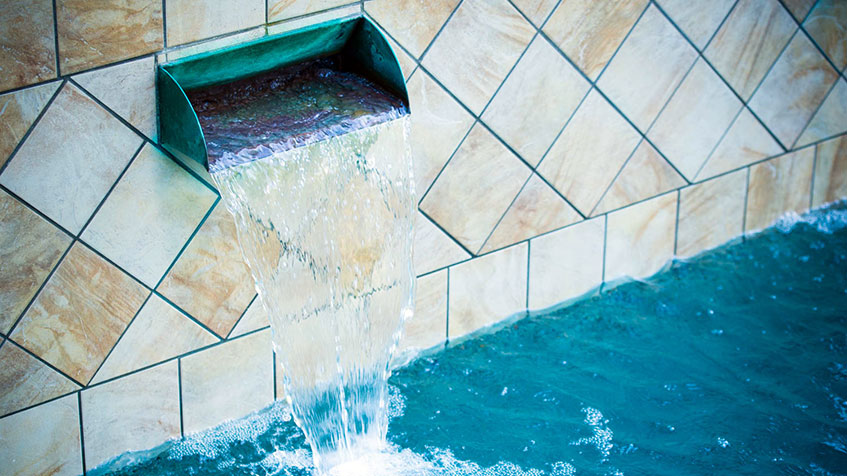 Water pours from a spout embedded in a tan tile wall into a clear pool.