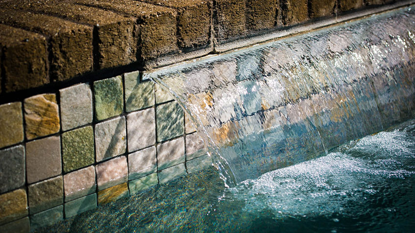 A sheet of water cascades over a mosaic tile wall into a pool beneath a brick ledge.