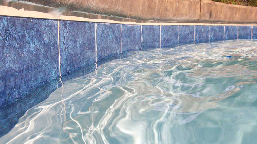 A row of deep blue tiles reflects shimmering water near a pool's coping edge.