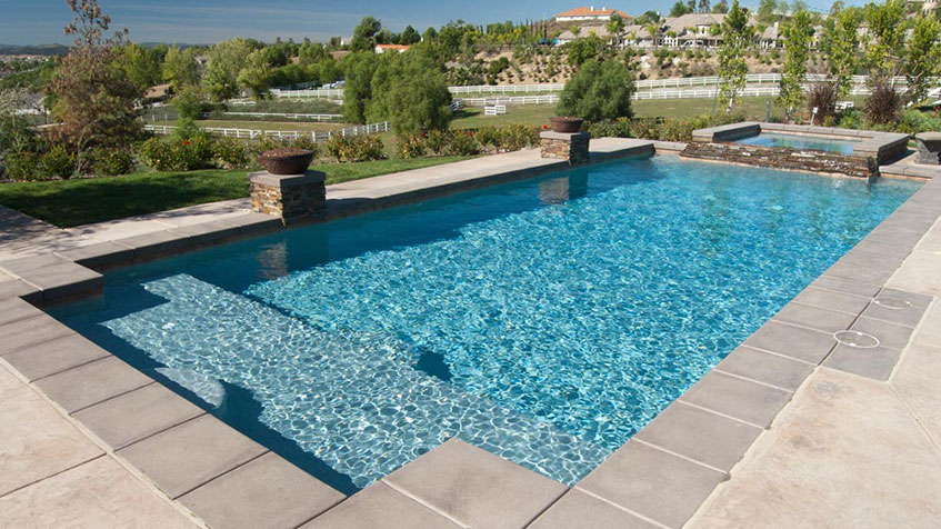 A rectangular backyard pool with a spa and wide entry steps overlooks a fenced hillside landscape.