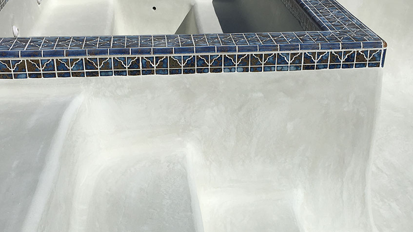 Close-up of a white pool edge with deteriorating plaster showing wear and staining, bordered by blue decorative tile trim at the waterline.